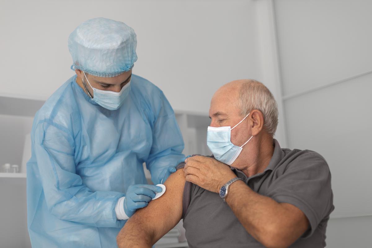 Hombre de 60 años que está por ser vacunado por un enfermero con traje y barbijo celeste en una habitación de paredes blancas